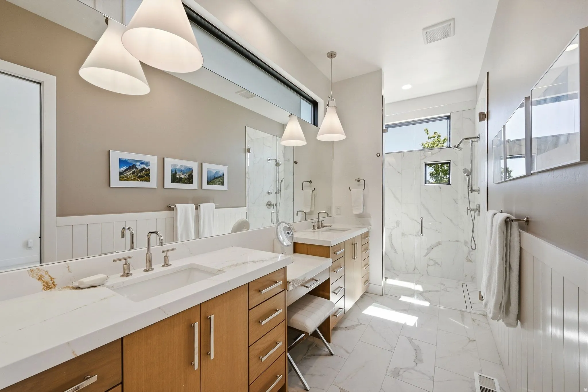 Spacious bathroom featuring a dual vanity with light wood cabinetry and white countertops, a separate vanity area, a walk-in shower with floor-to-ceiling tile, and modern pendant lighting