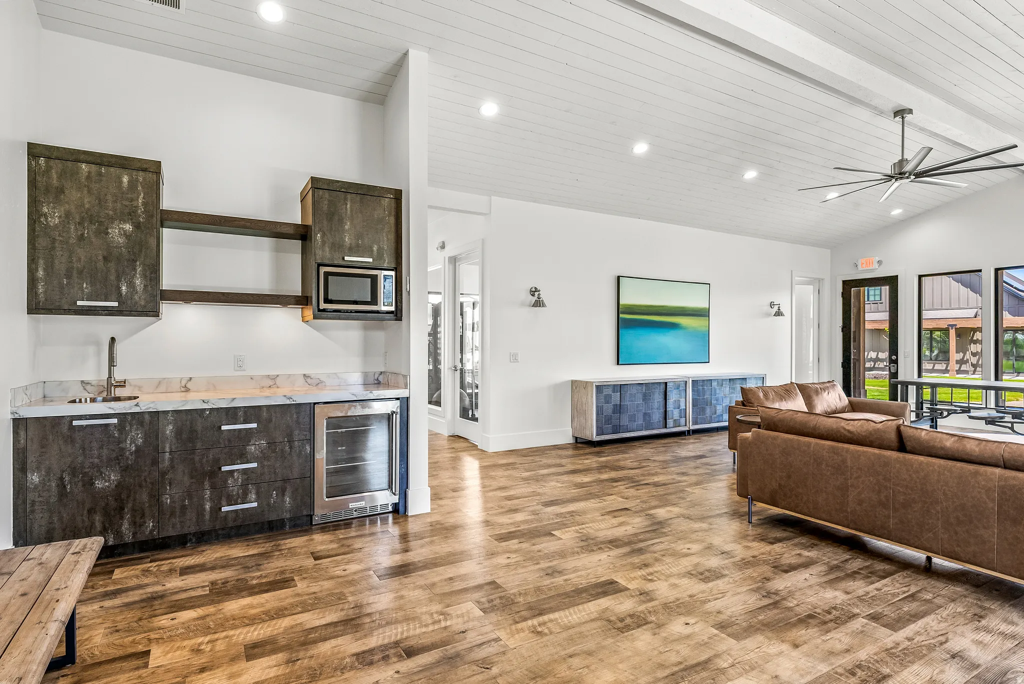 Spacious open-concept room featuring a built-in wet bar with a microwave and beverage refrigerator, wood-finish flooring, vaulted shiplap ceiling with recessed lighting and a ceiling fan, and large windows