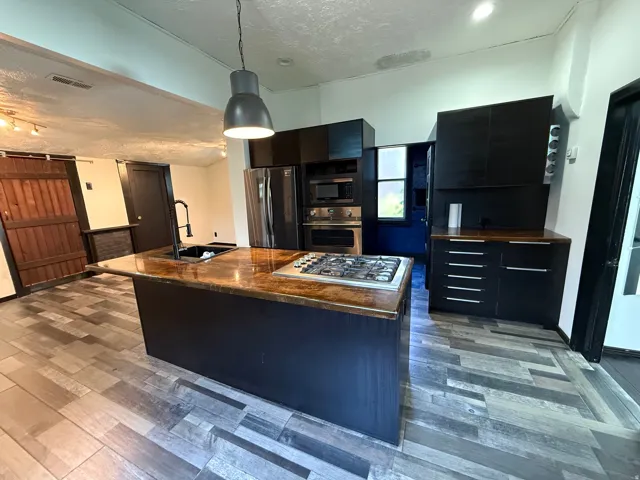 Wood-finish flooring, central island with integrated gas cooktop, black cabinetry, stainless steel appliances, and a modern pendant light fixture