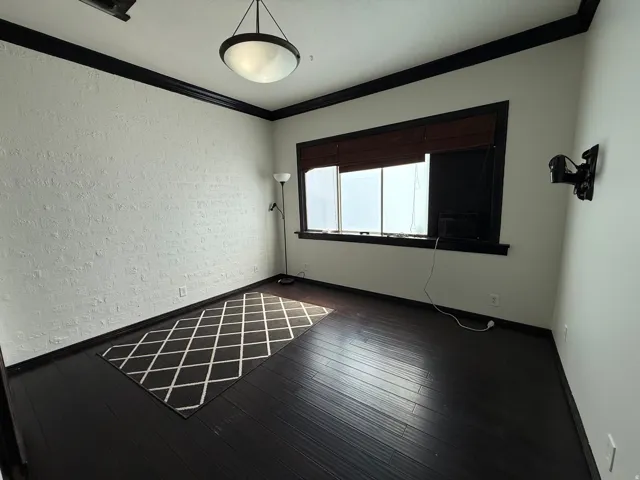 Dark-finish wood flooring, white painted brick accent wall, contrasting dark trim, window with blinds, and ceiling-mounted light fixture
