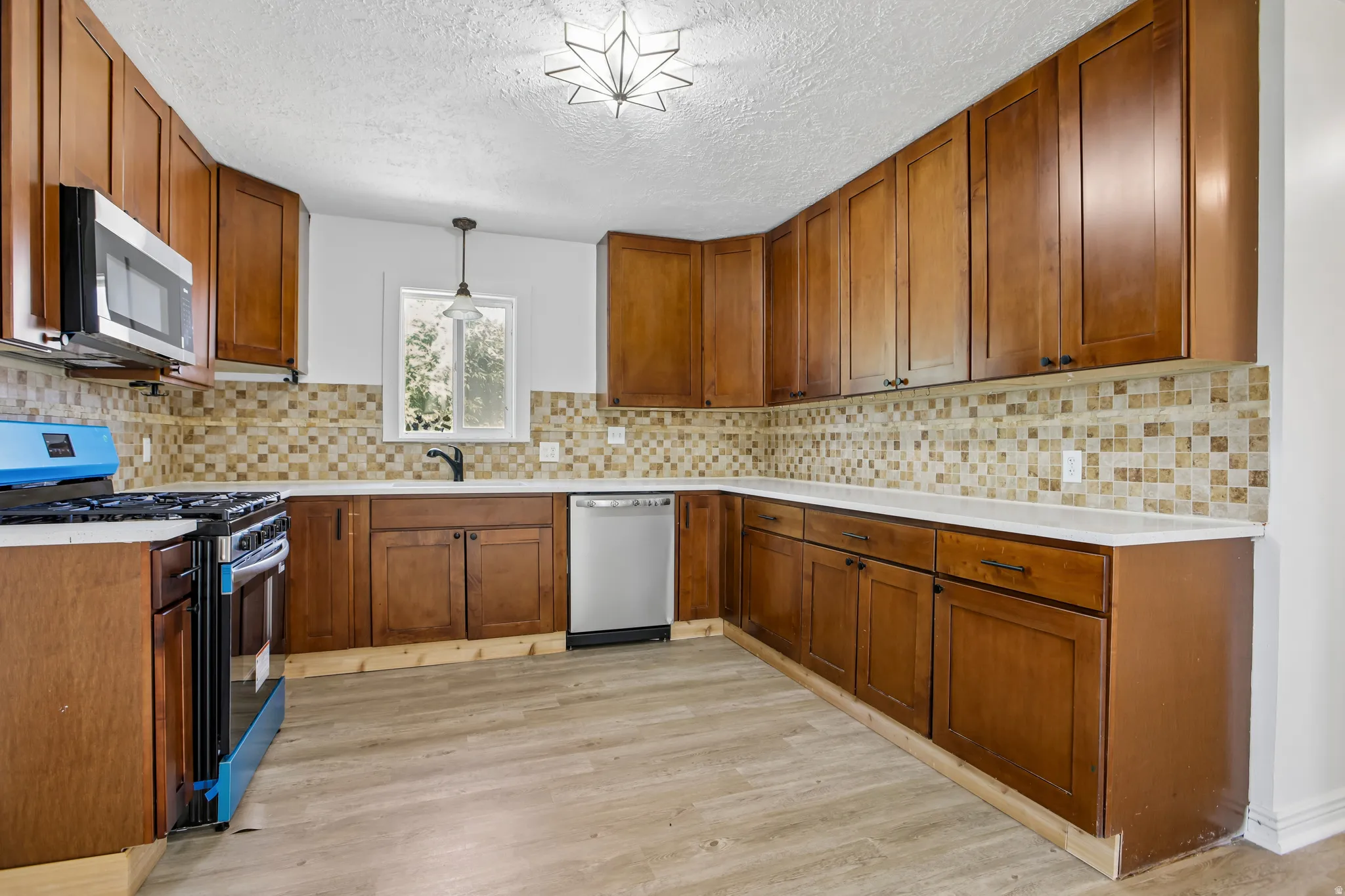 Kitchen featuring wood-finish cabinetry, light-toned countertops, a mosaic tile backsplash, wood-finish flooring, and a stainless steel over-the-range microwave