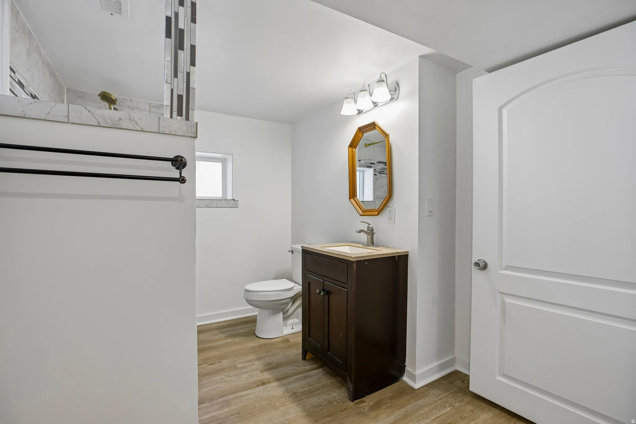 Bathroom featuring wood-finish flooring, a dark wood vanity with a light-toned countertop, and a decorative gold-framed mirror