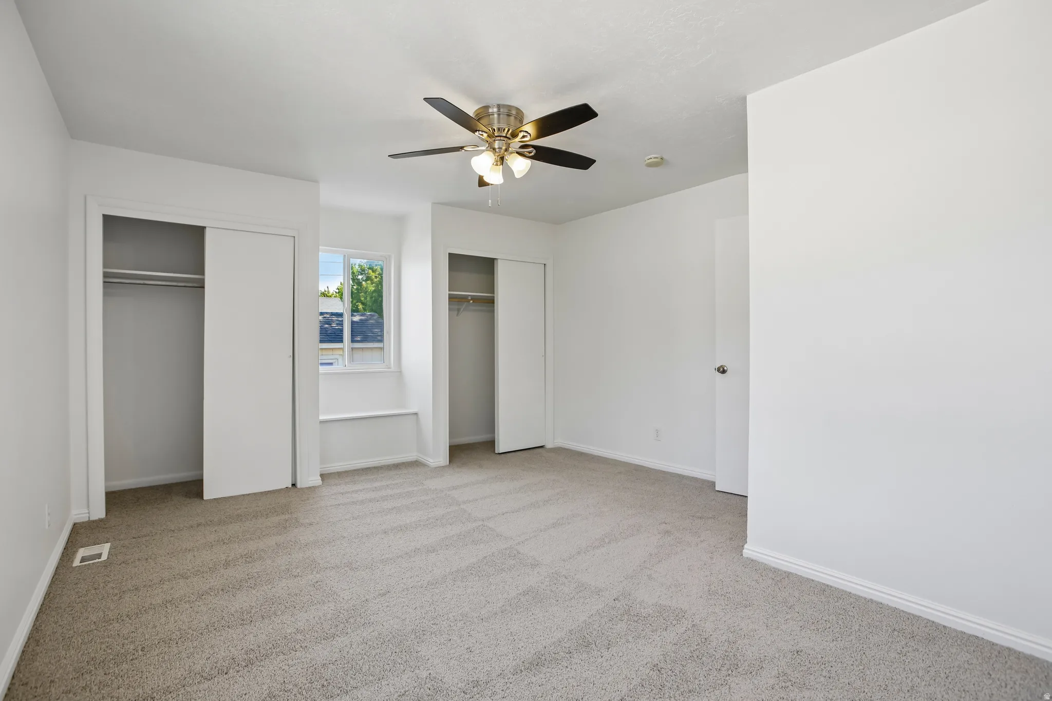 Spacious room featuring neutral carpeting, a ceiling fan with integrated lighting, two sliding door closets with hanging rods, and a window with a built-in ledge