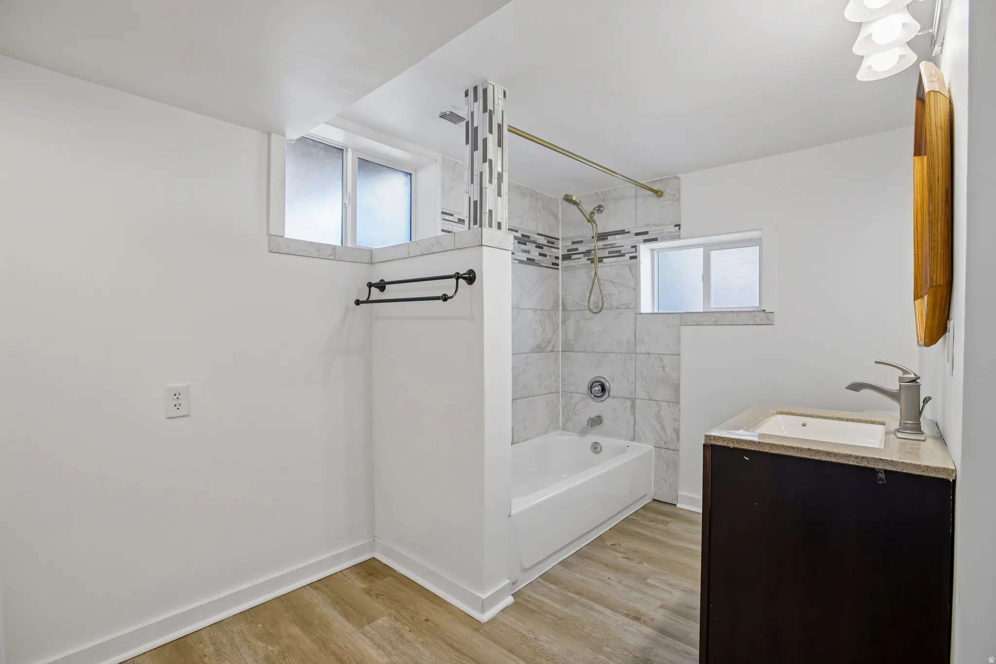 Bathroom featuring a white bathtub with tile surround, wood-finish flooring, a dark wood-finish vanity with a light-colored countertop and integrated sink, and frosted windows