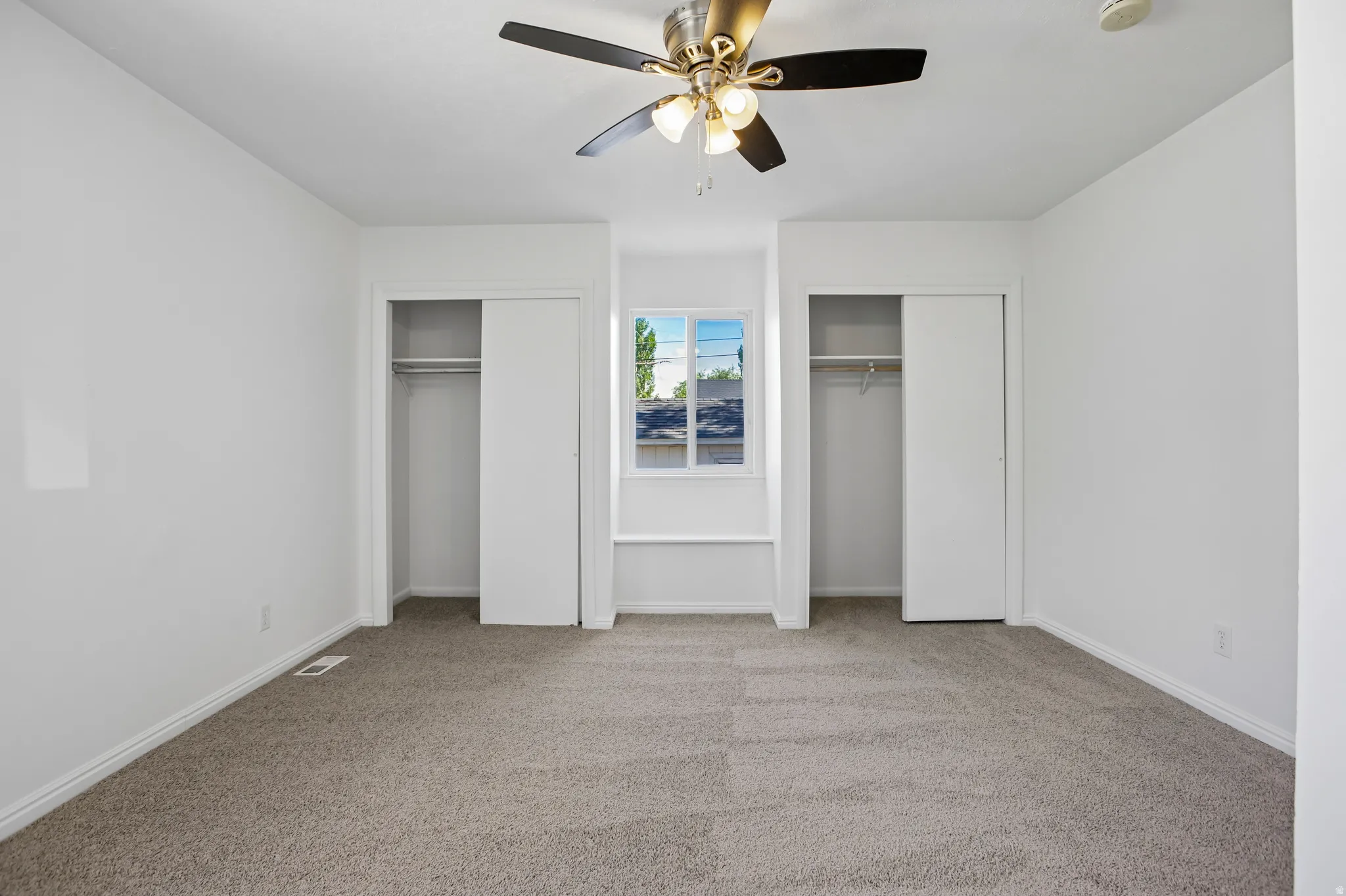 Carpeted room featuring a ceiling fan with integrated lighting, two sliding door closets, and a central window with a built-in window seat