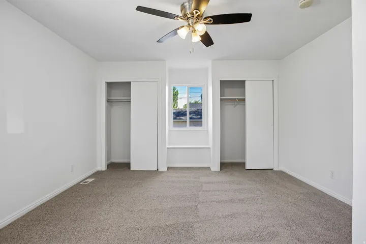 Carpeted room featuring a ceiling fan with integrated lighting, two sliding door closets, and a central window with a built-in window seat