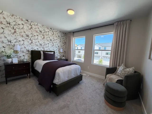 Bedroom featuring a floral accent wall, two large windows, neutral-toned curtains, and gray carpeting