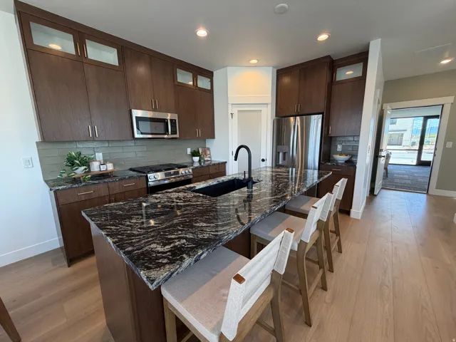Contemporary kitchen featuring dark wood cabinetry, stainless steel appliances, a black granite island with an undermount sink, and light wood-finish flooring