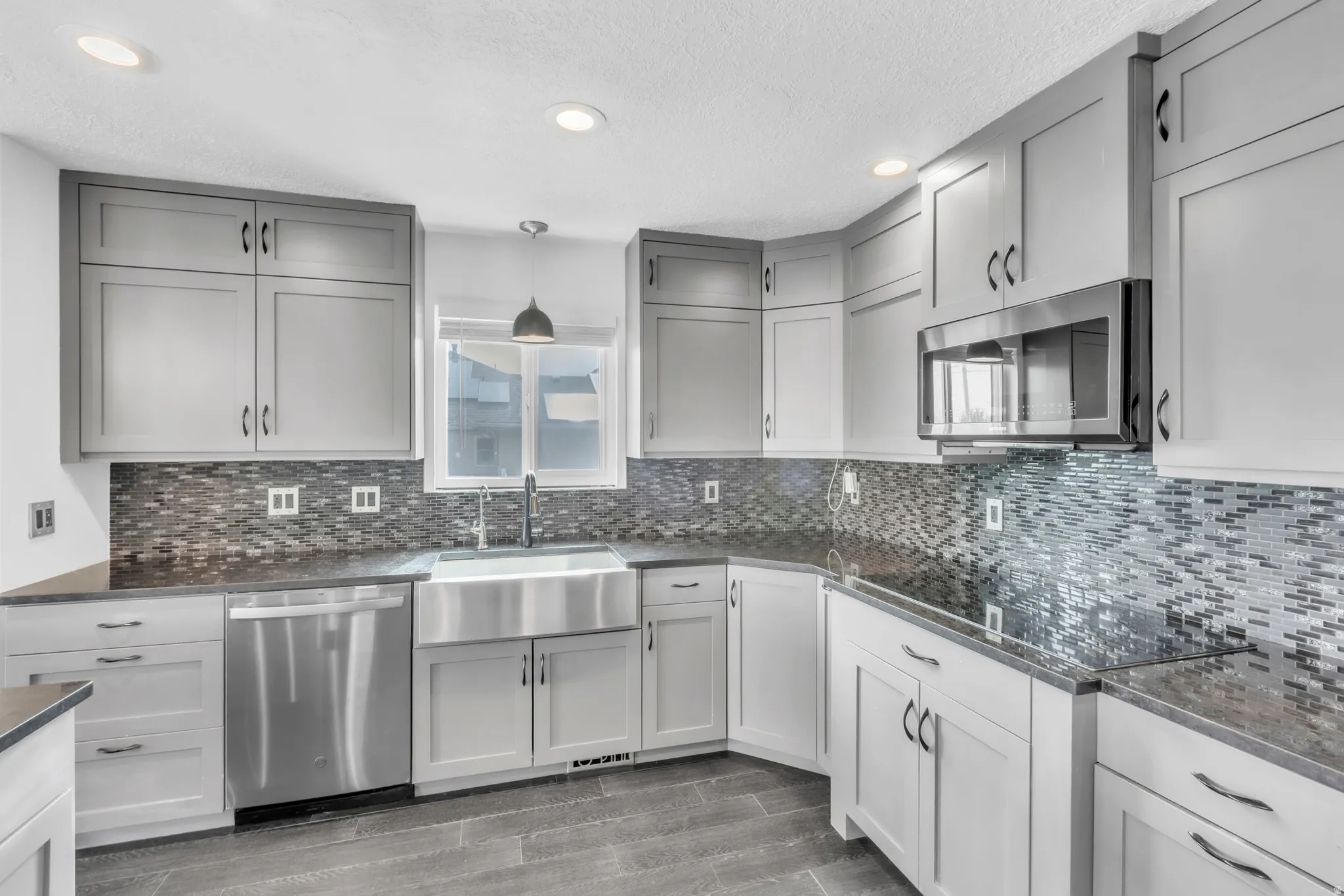 Contemporary kitchen featuring light gray shaker cabinetry, dark countertops, a stainless steel apron sink, mosaic tile backsplash, and stainless steel appliances