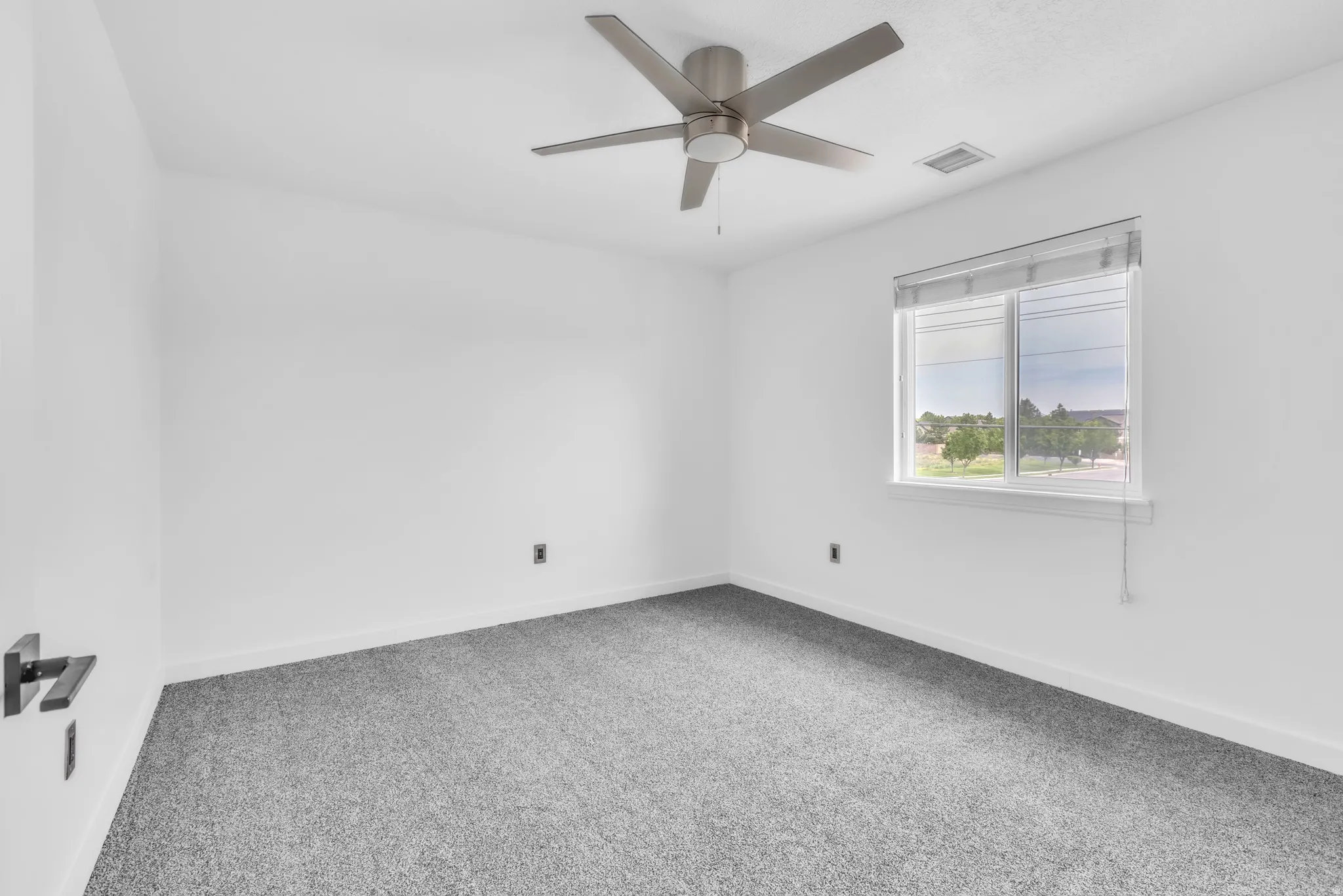 Neutral-toned room featuring a large window with blinds, ceiling fan with brushed nickel accents, and plush grey carpeting