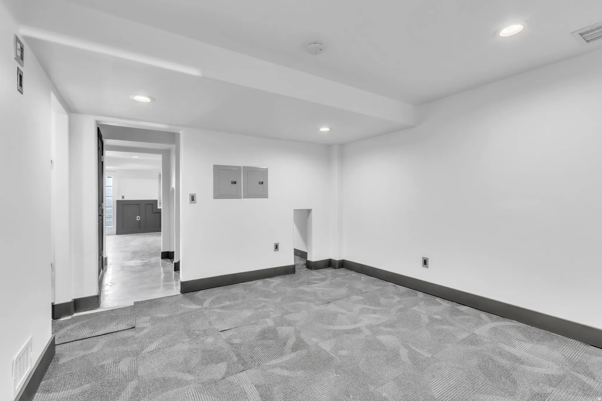 Spacious room featuring gray pattern carpet, recessed ceiling lighting, crisp white walls, dark gray baseboards, and a doorway leading to an additional space