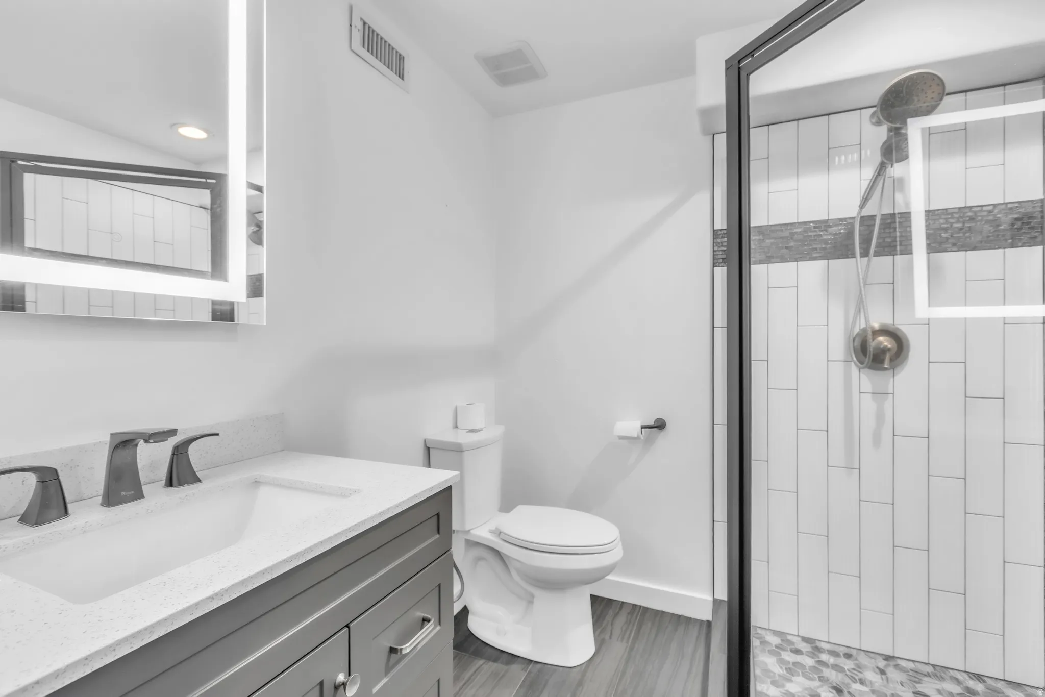 Bathroom featuring a gray vanity with an integrated sink and quartz countertop, an illuminated mirror, a glass-enclosed shower with subway tile and a mosaic accent, and wood-finish flooring