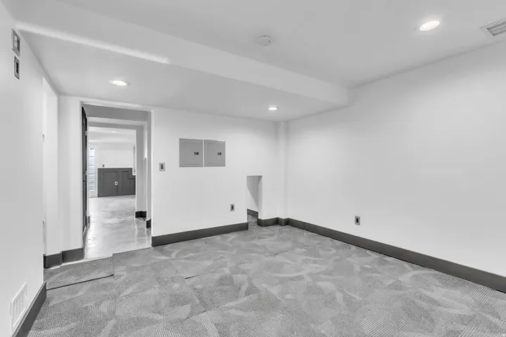 Spacious room featuring gray pattern carpet, recessed ceiling lighting, crisp white walls, dark gray baseboards, and a doorway leading to an additional space