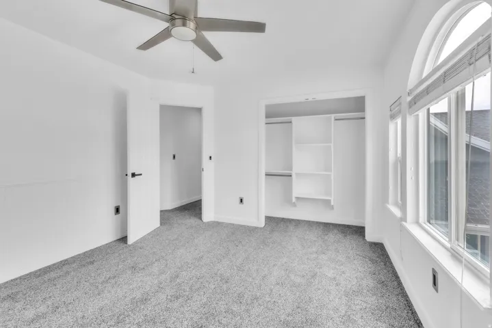 Bedroom featuring a ceiling fan with brushed nickel finish, plush gray carpet, a built-in closet with shelving, and an arched window with blinds