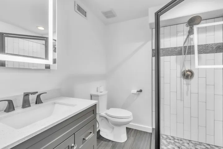 Bathroom featuring a gray vanity with an integrated sink and quartz countertop, an illuminated mirror, a glass-enclosed shower with subway tile and a mosaic accent, and wood-finish flooring