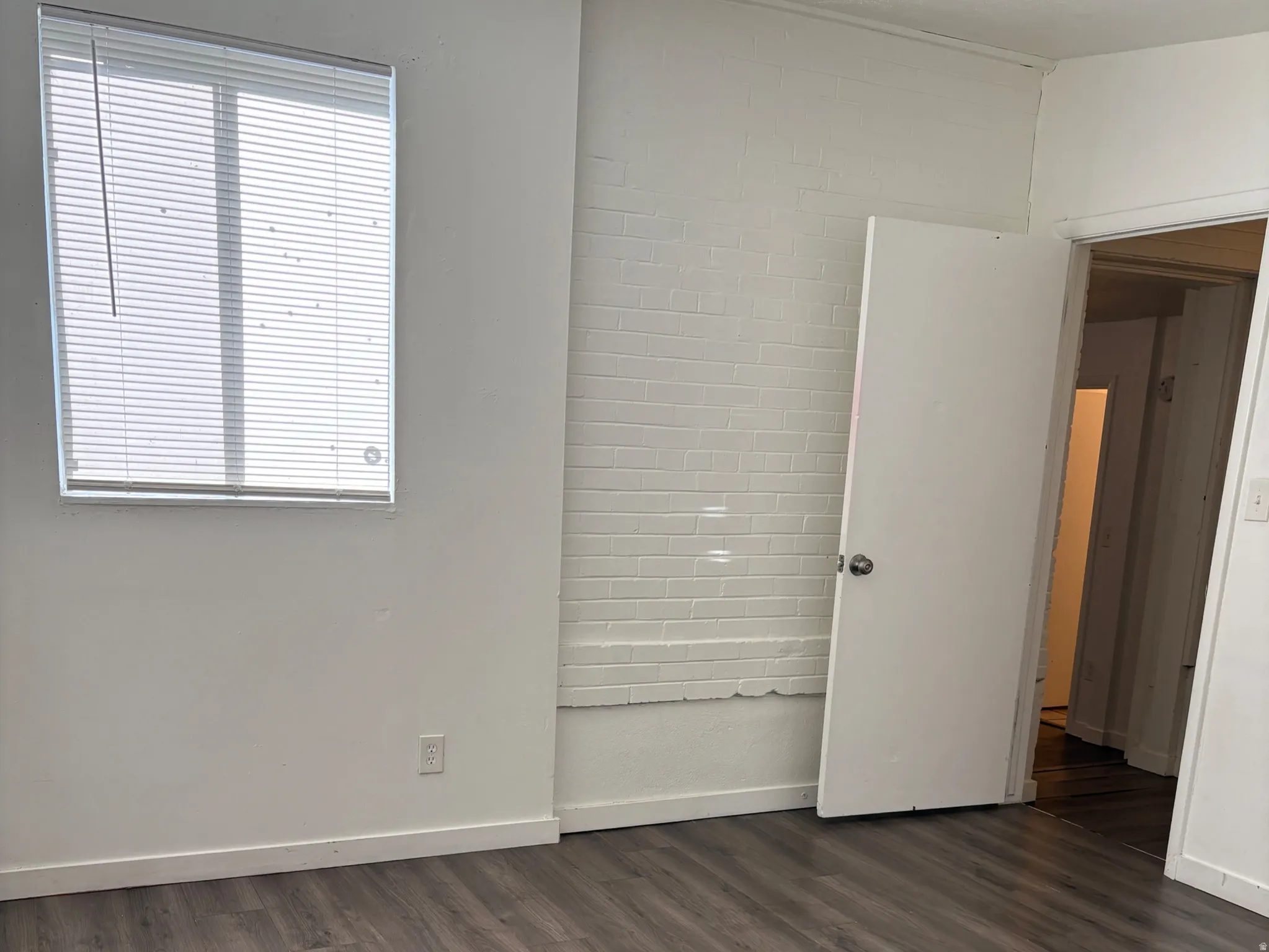 Spacious interior featuring wood-finish flooring, a bright window with blinds, and a painted brick accent wall