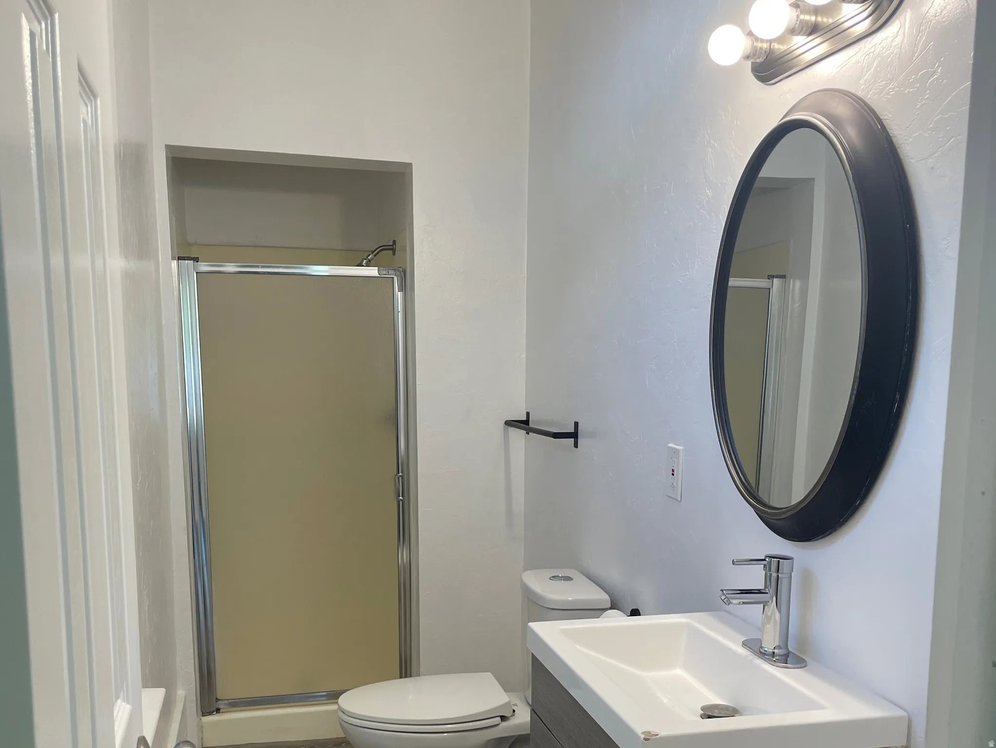 Compact bathroom featuring a shower enclosure, contemporary vanity with integrated sink, wood-finish flooring, toilet, and an oval wall mirror