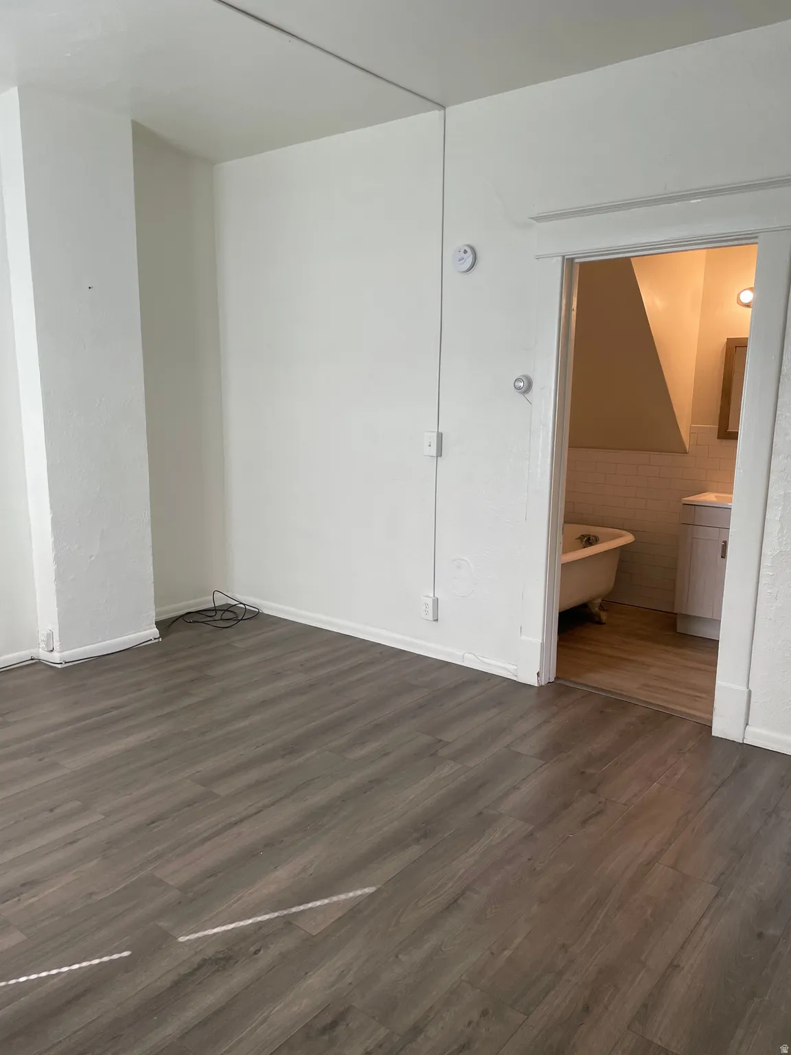 Spacious interior room featuring wood-finish flooring, white baseboards, and a doorway opening to a bathroom with a clawfoot tub and tile wainscoting