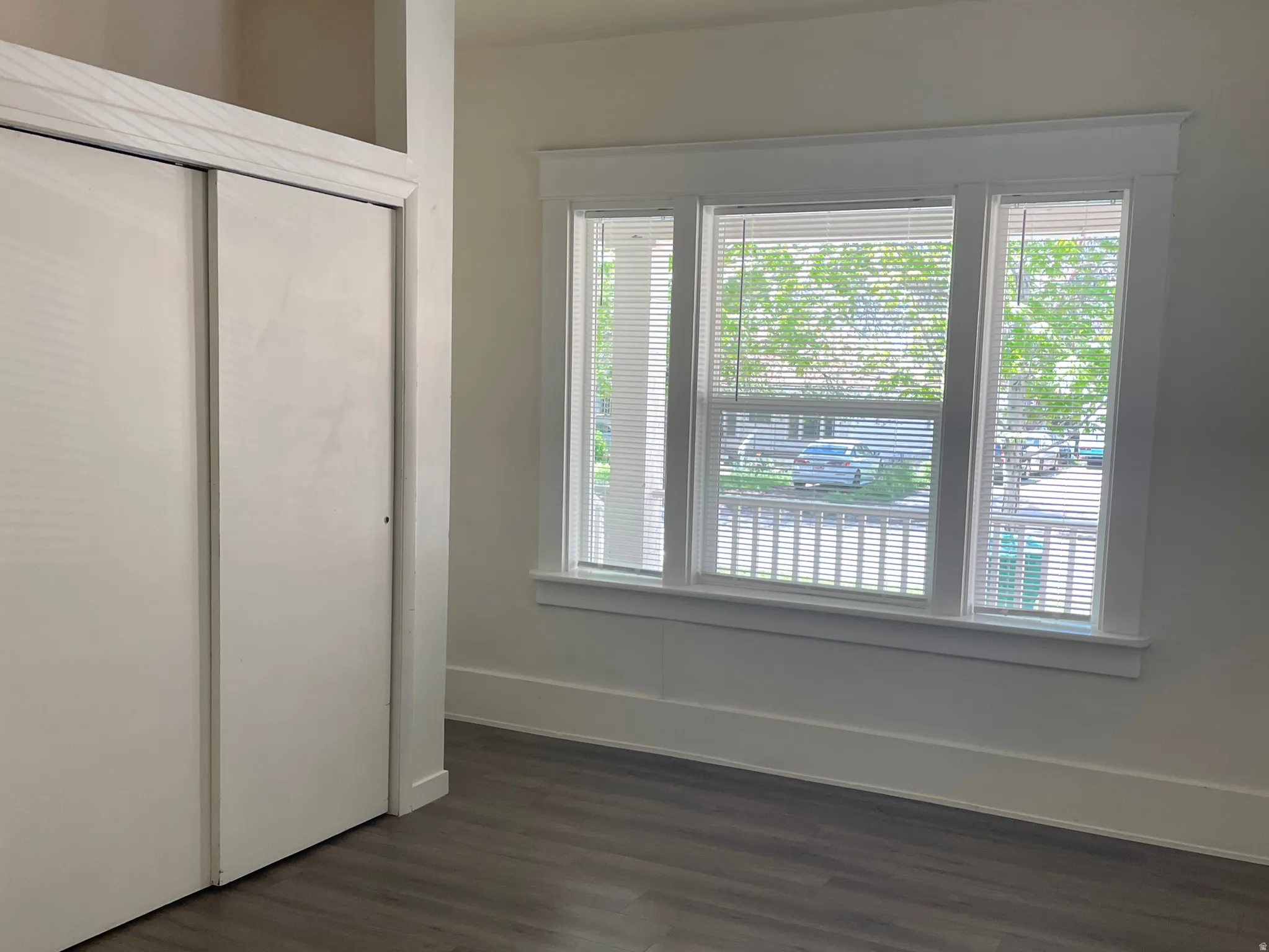 Bright room featuring wood-finish flooring, a large window with white trim, white horizontal blinds, and a built-in closet with white sliding doors and an upper storage shelf