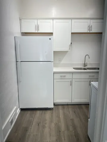 Compact kitchen area featuring white cabinetry, a stainless steel sink with a single-handle faucet, a white refrigerator, and wood-finish flooring