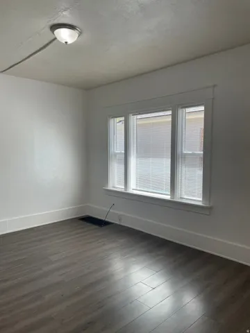 Room featuring large triple window with blinds, wood-finish flooring, white walls, and white trim