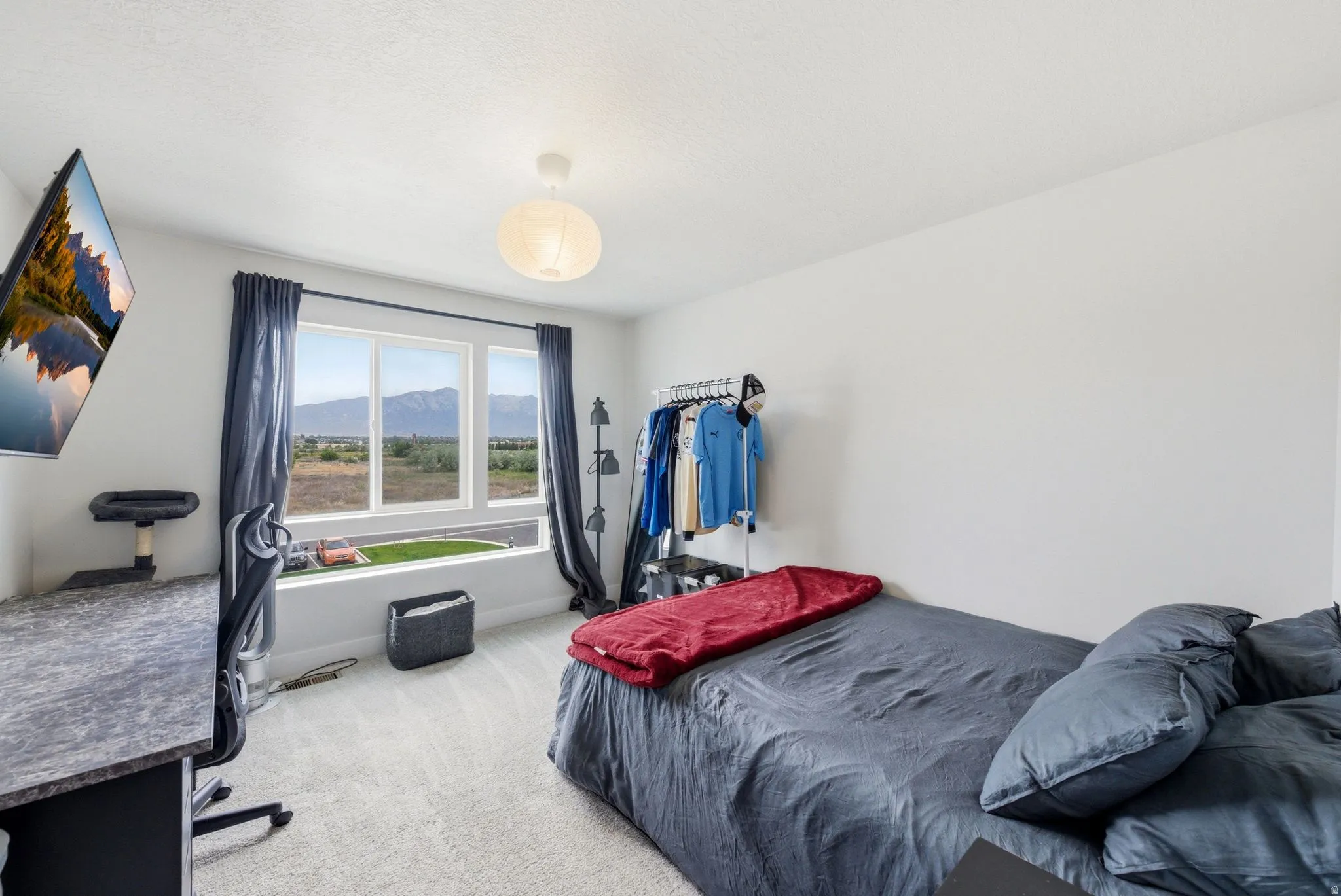 Light-filled room featuring a large window with mountain scenery, plush carpeting, a modern ceiling light fixture, a built-in wall-mounted television, and a desk with a textured surface