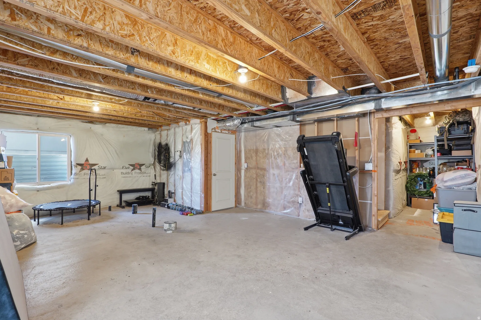 Unfinished basement space featuring exposed wood joists and subflooring, a white door, and a single window with horizontal blinds