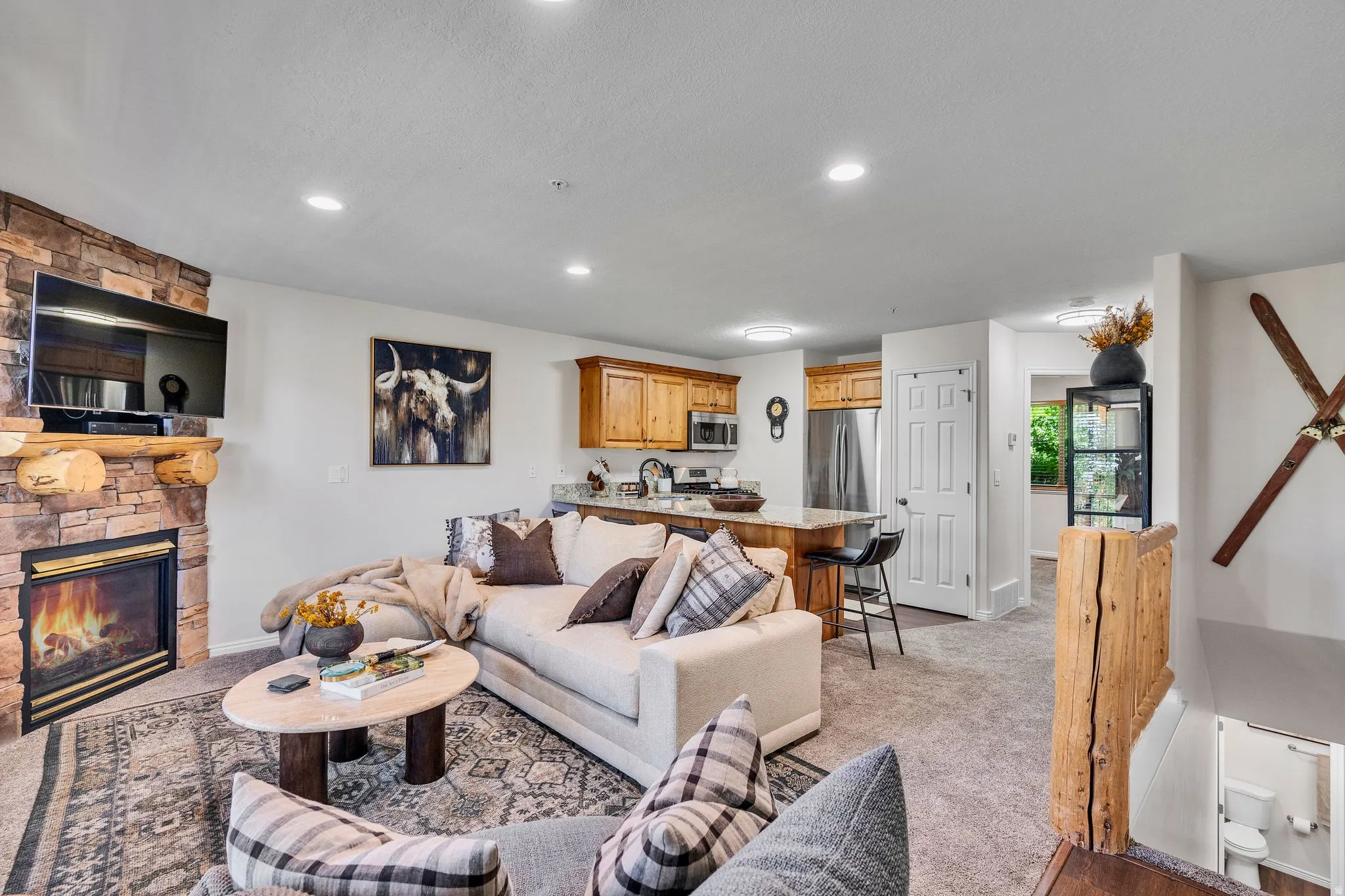 Open-concept living space featuring a stacked stone fireplace with a log mantel, recessed lighting, and soft gray carpeting