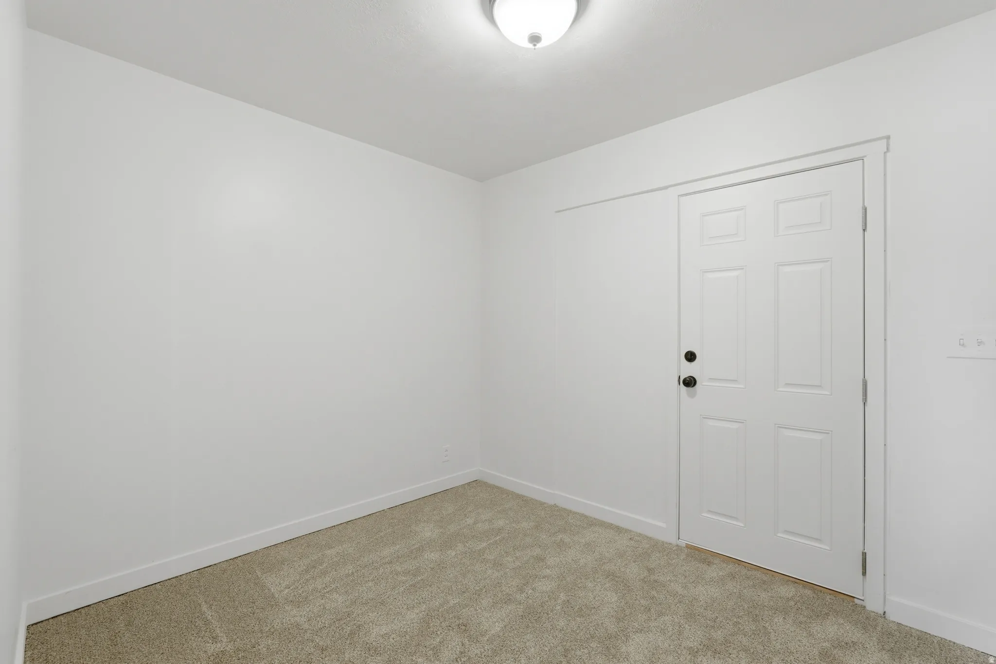 Room featuring light-toned carpeting, white walls, and a white six-panel door with matte black hardware