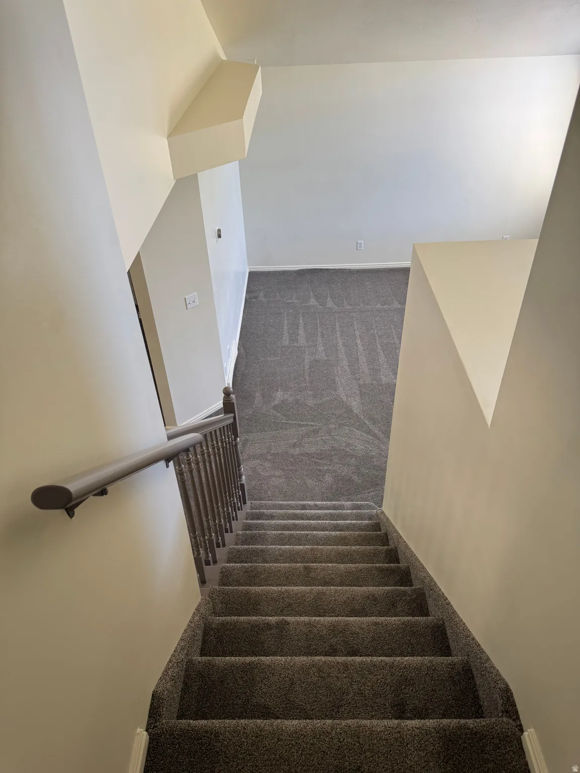 Carpeted staircase with a wood-finish handrail and balusters, leading to a lower level featuring wall-to-wall carpeting and neutral-toned walls