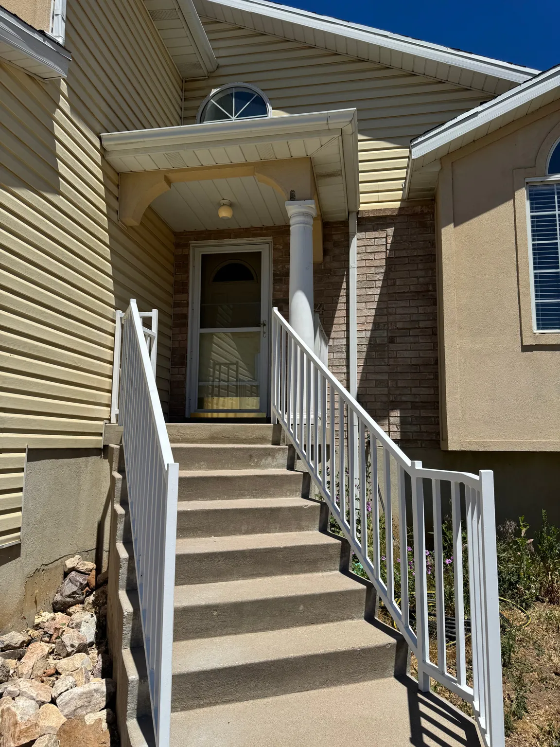 Inviting entryway featuring a covered porch, durable concrete steps, white metal railings, a half-round transom window, and a combination of siding and brick facade
