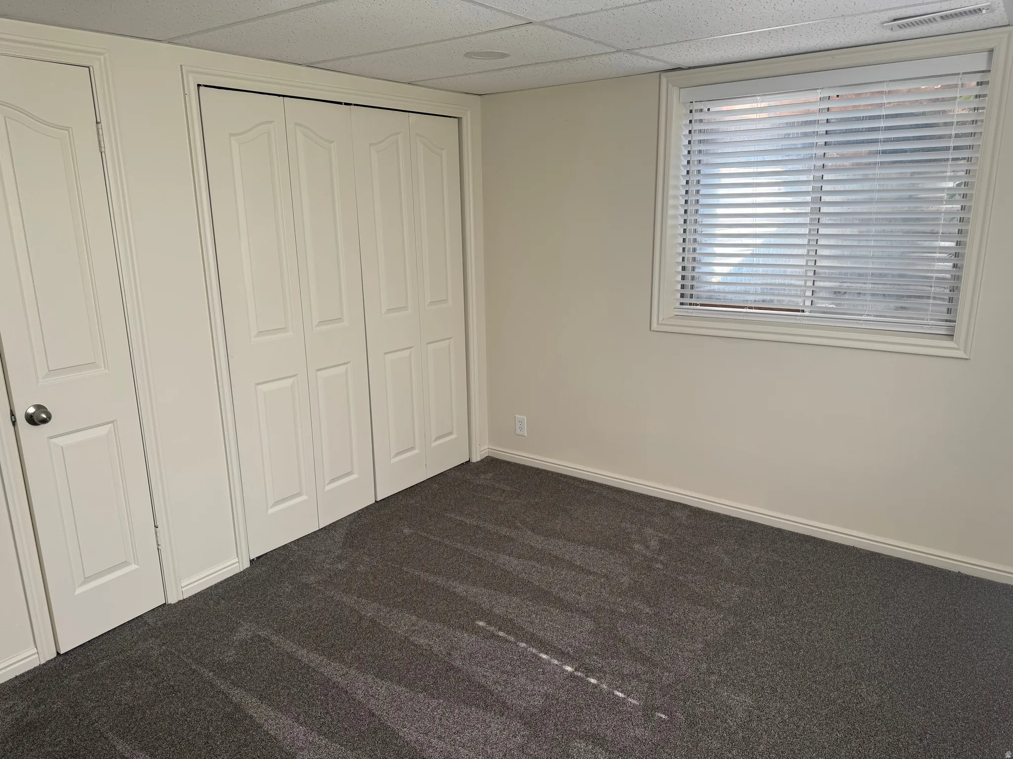 Neutral-toned interior featuring grey carpet flooring, a window with horizontal blinds, and paneled bifold closet doors