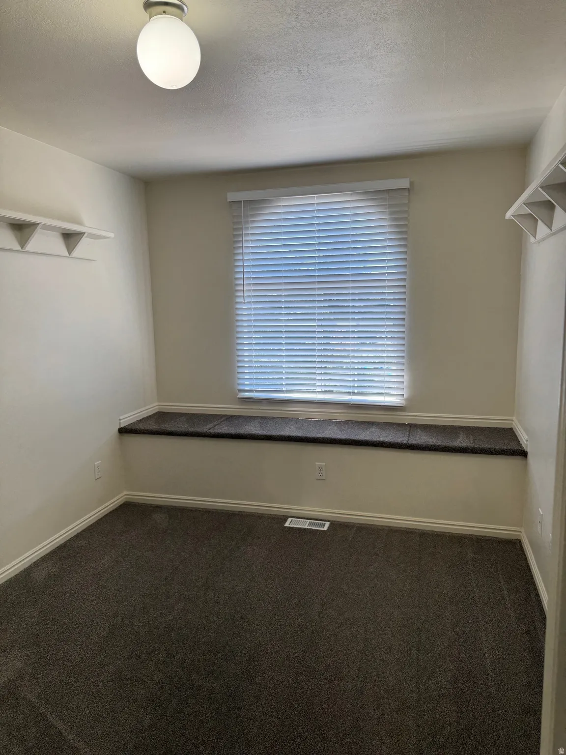 Room featuring a window with blinds, a built-in window seat, dark grey carpeting, a ceiling-mounted light fixture, and white wall shelving