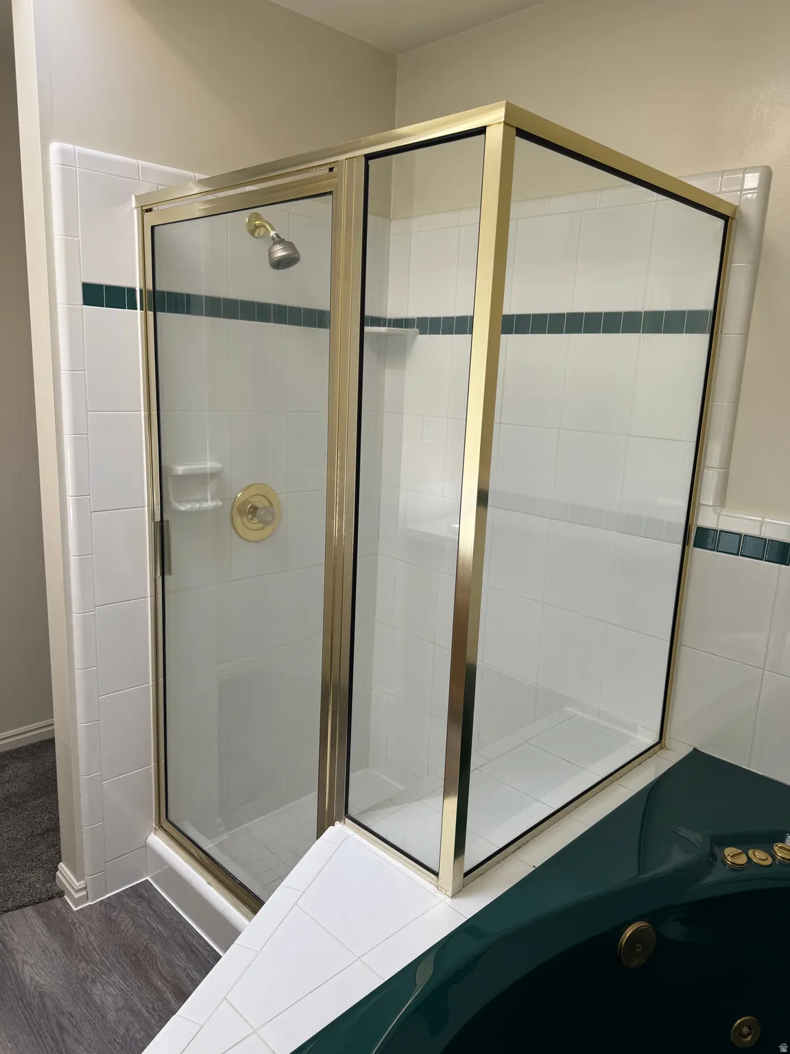 Gold-framed glass shower enclosure with a built-in shelf, white tile walls, a green accent tile band, and a coordinating green corner tub with brass fixtures