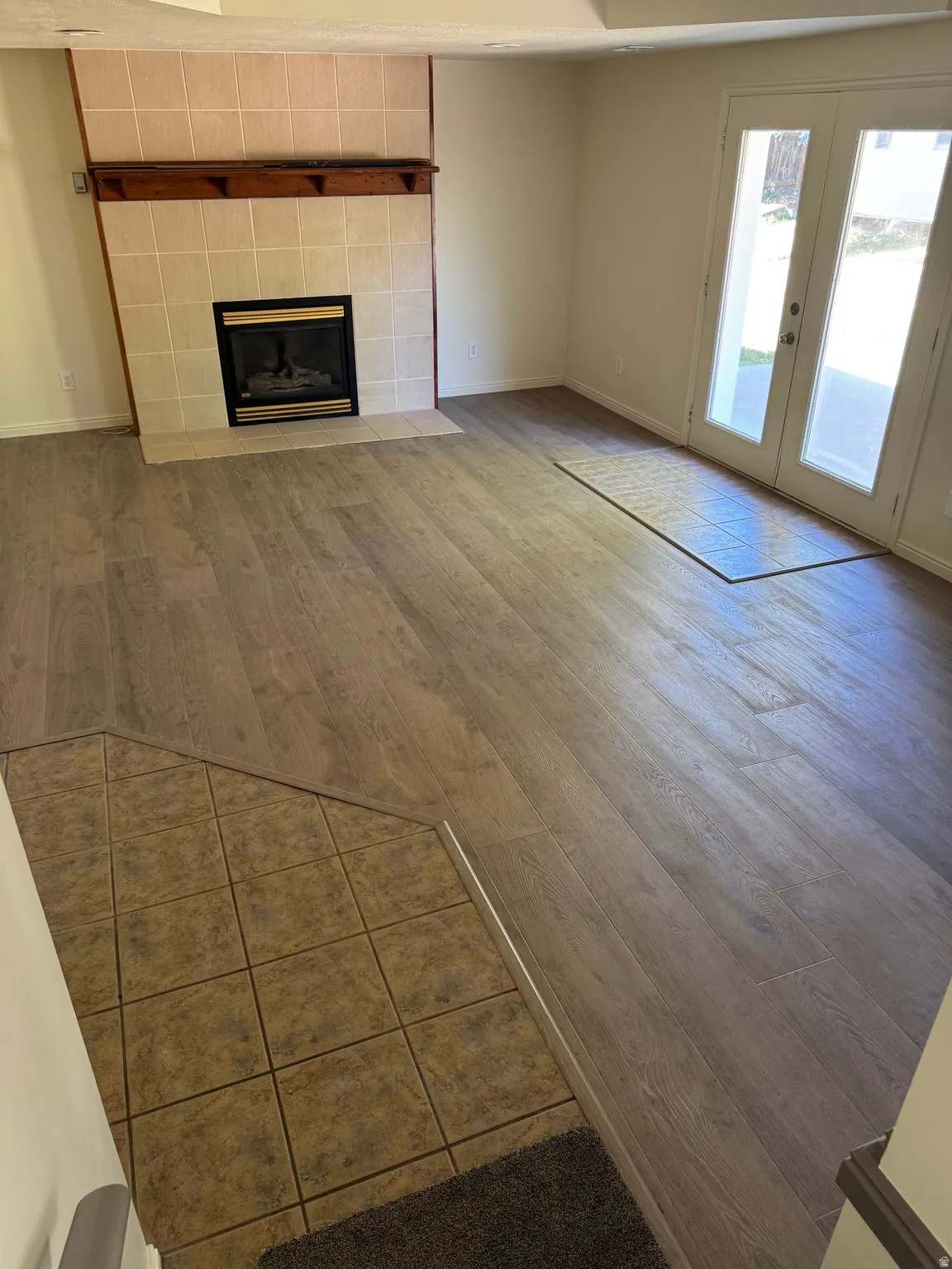 Spacious living area featuring a tiled fireplace with a wood mantle, wood-finish flooring, and double glass-paneled doors leading to an outdoor patio