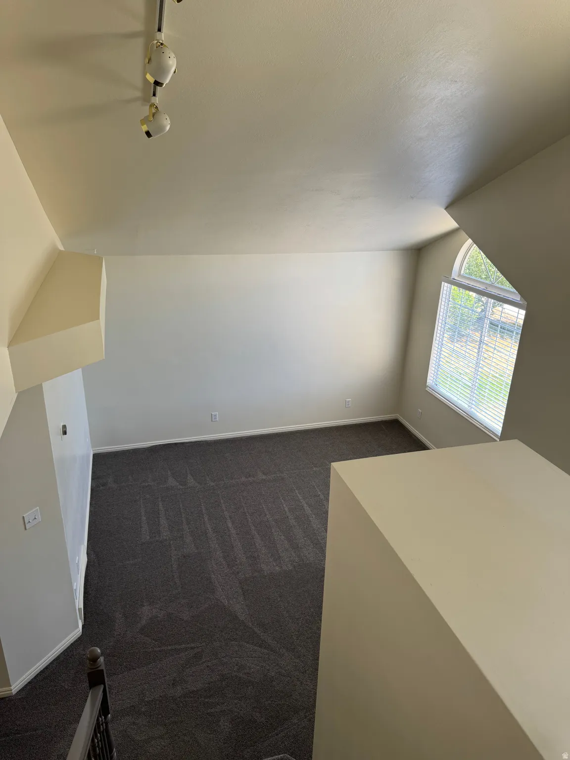 Vaulted ceilings with track lighting illuminate this carpeted room featuring a large arched window with blinds