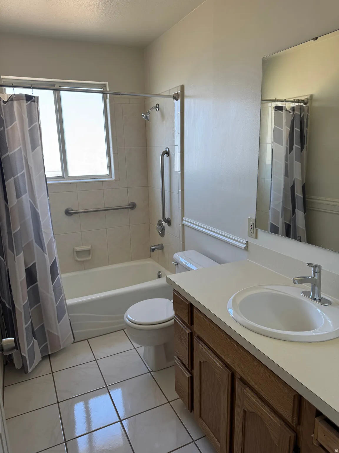 Bathroom featuring a white bathtub with grab bars, a window, a single-sink vanity with wood-finish cabinetry, a toilet, and tiled flooring