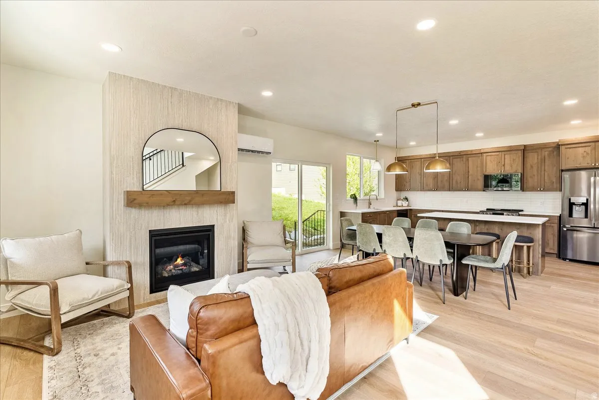 Open-concept living space featuring a fireplace with a wood mantel, light wood-finish flooring, recessed lighting, and a kitchen with wood cabinetry, stainless steel appliances, and a white subway tile backsplash