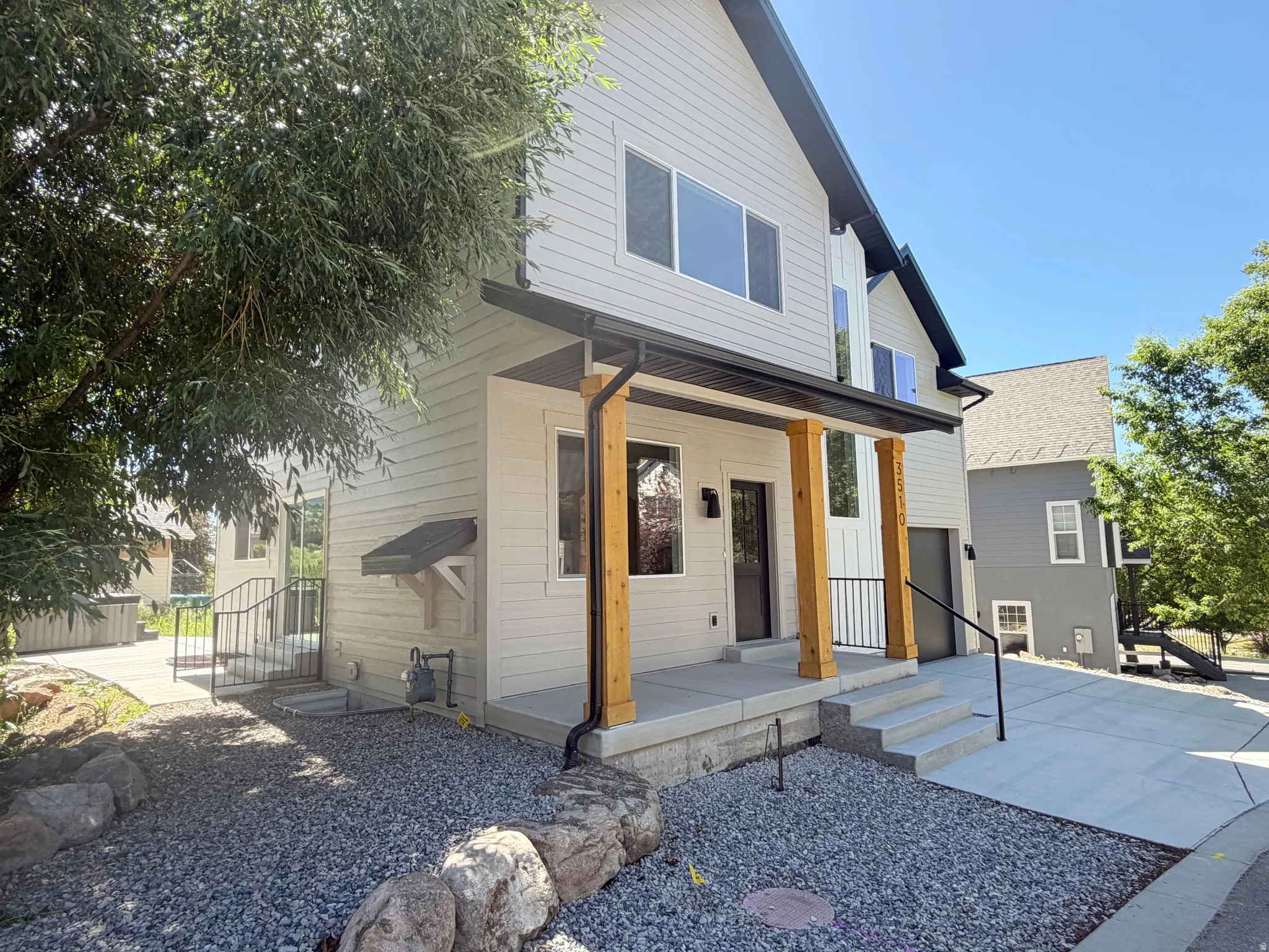 Modern two-story residence featuring light-colored siding, a covered front porch with wood columns, dark trim, and a gravel front yard with rock landscaping