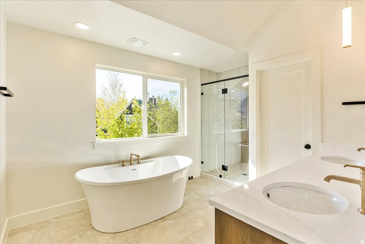 Spacious bathroom featuring a freestanding soaking tub with brass fixtures, a glass-enclosed shower, dual vanity with undermount sinks and brass faucets, and tile flooring