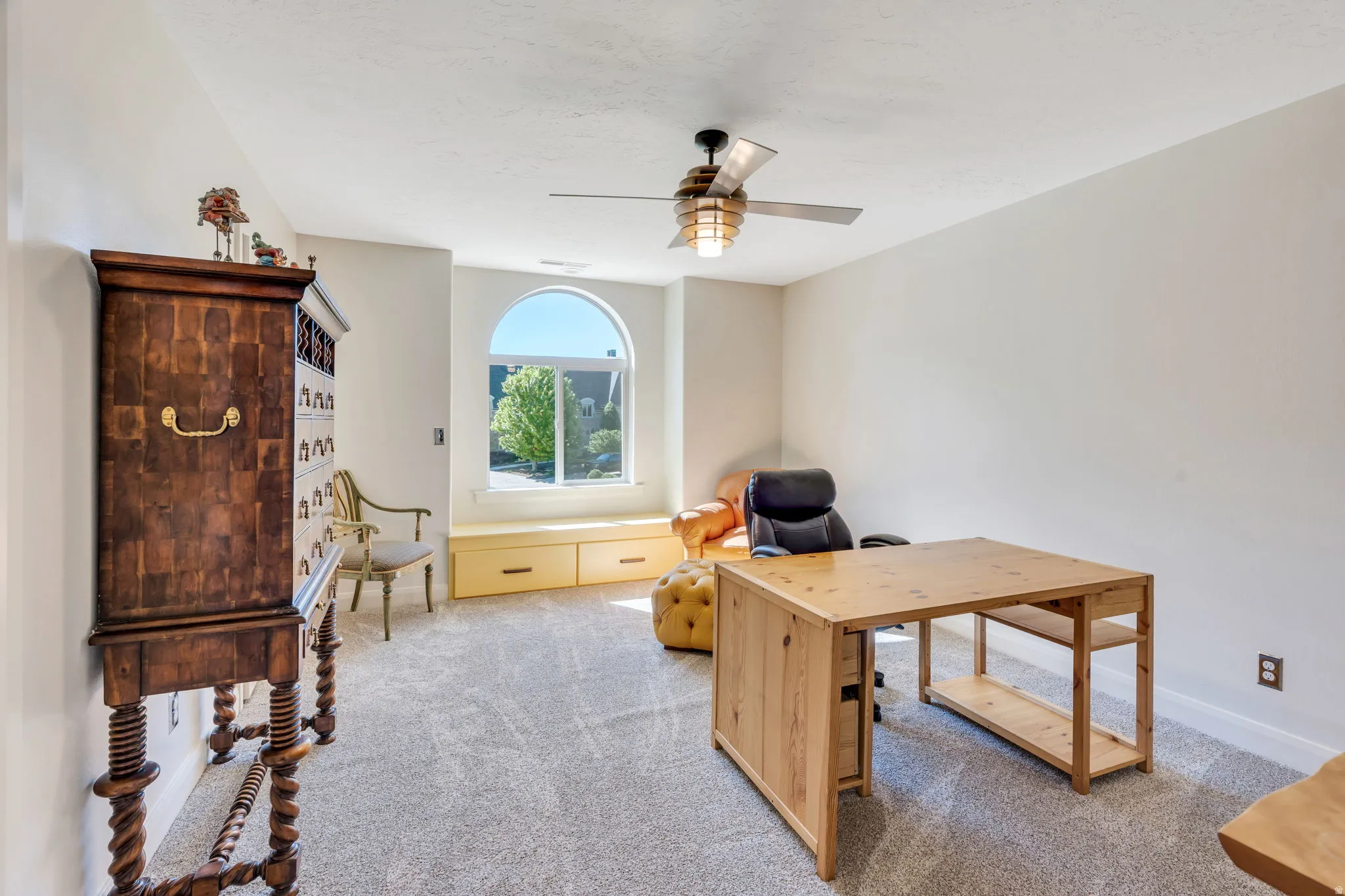 Spacious room featuring a large arched window with built-in window seating and storage, textured carpeting, and a ceiling fan with integrated lighting