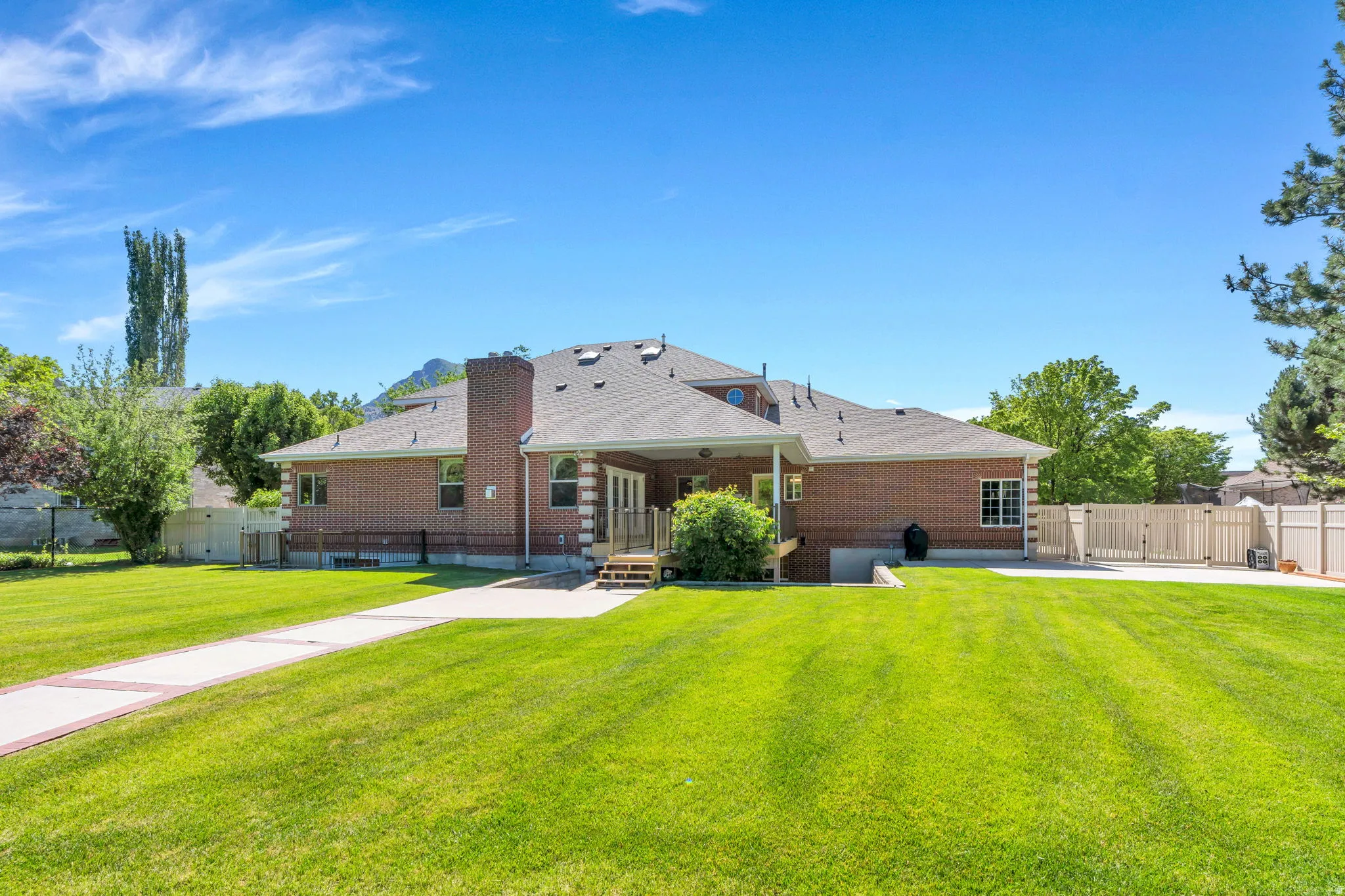 Expansive rear exterior featuring a red brick facade, a covered patio with wooden steps, a concrete pad, a manicured lawn, and a light-toned privacy fence