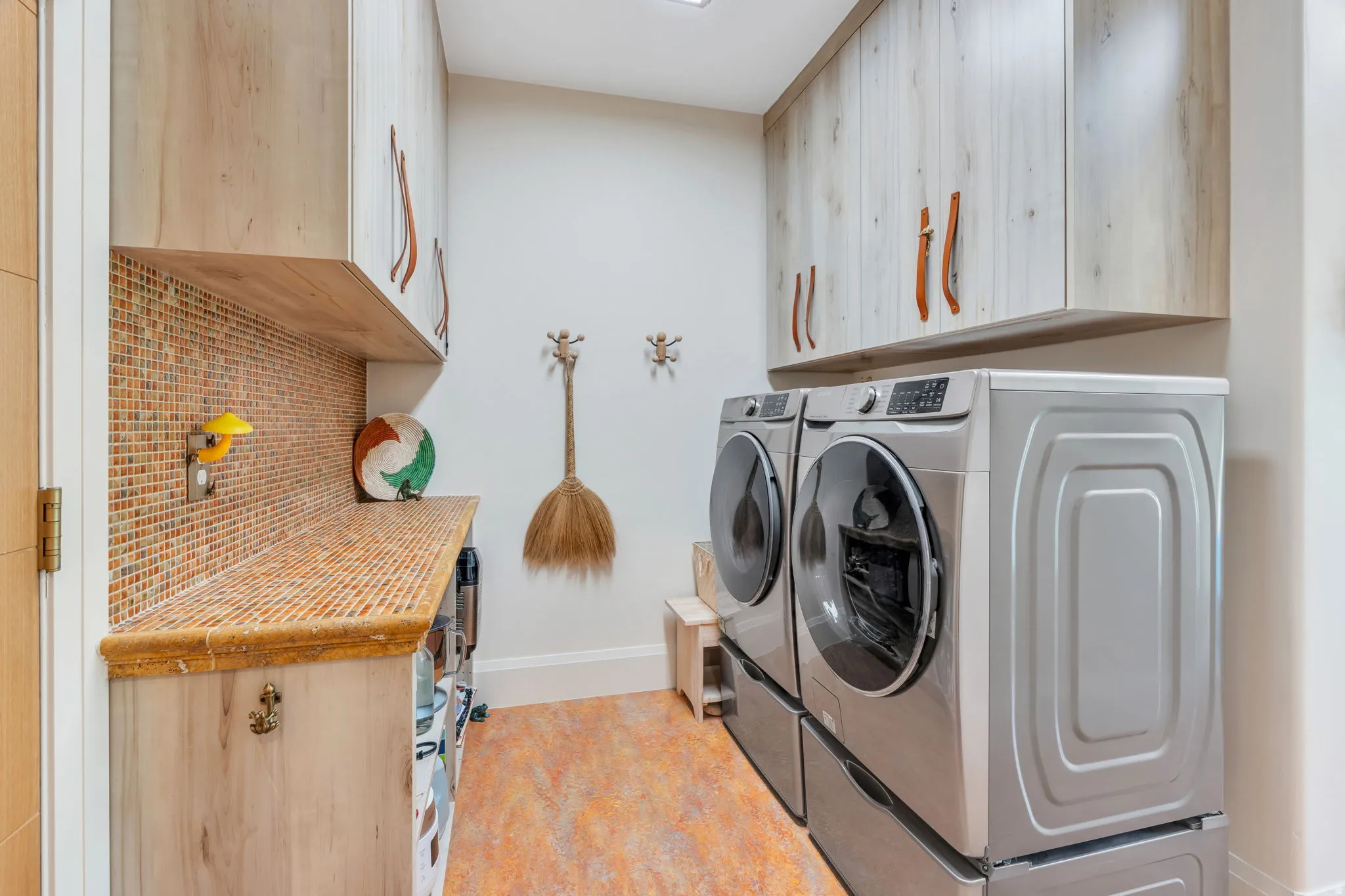 Laundry area featuring wood-finish cabinetry with leather-style pulls, a mosaic tile backsplash, and a coordinating countertop