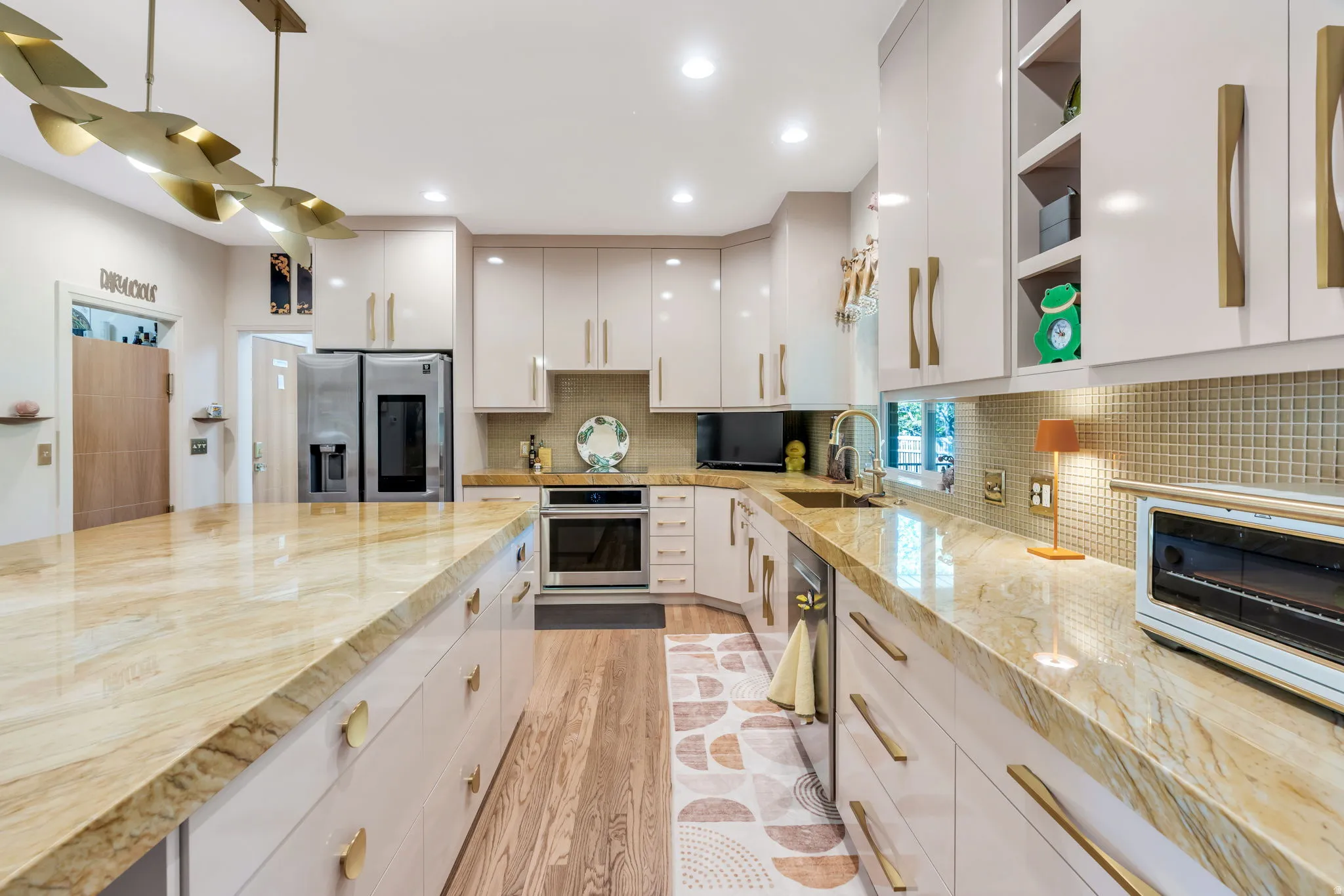 Spacious kitchen featuring extensive stone countertops, light-toned cabinetry with gold-toned hardware, wood-finish flooring, recessed lighting, and a built-in oven