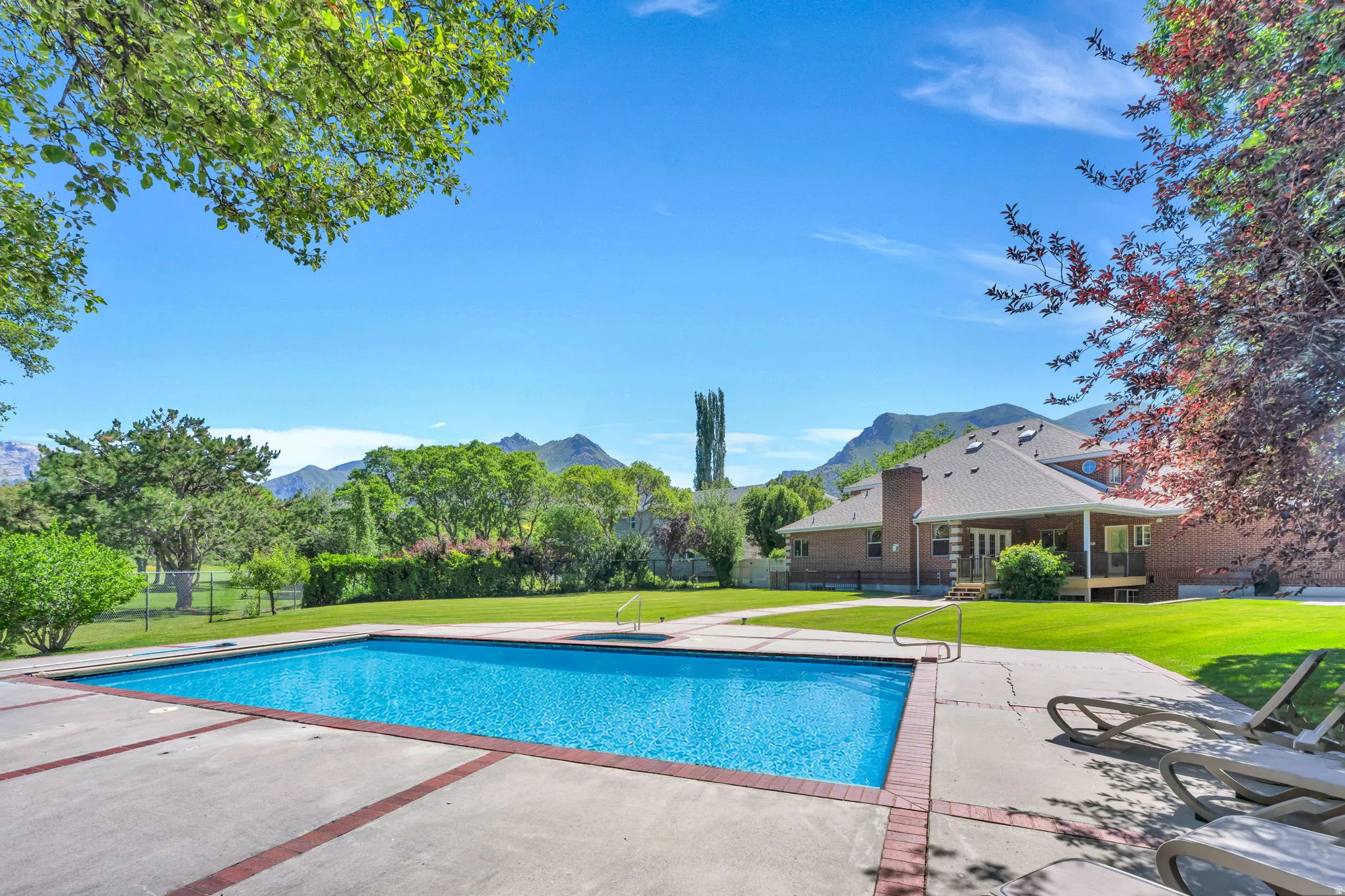 In-ground swimming pool featuring a concrete surround with brick accents, extensive green lawn, and mature trees