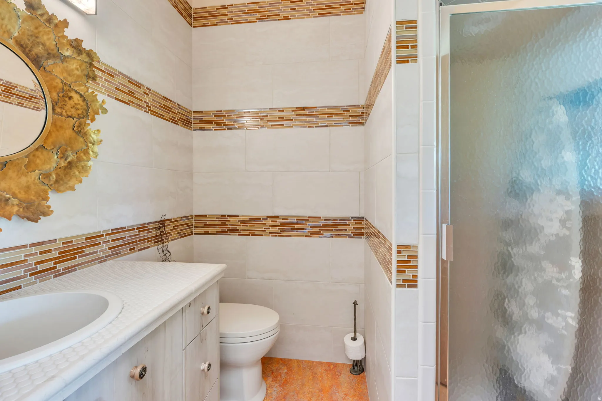 Bathroom featuring a single vanity with an integrated sink, white tile walls with decorative mosaic accents, a toilet, and a frosted glass shower enclosure