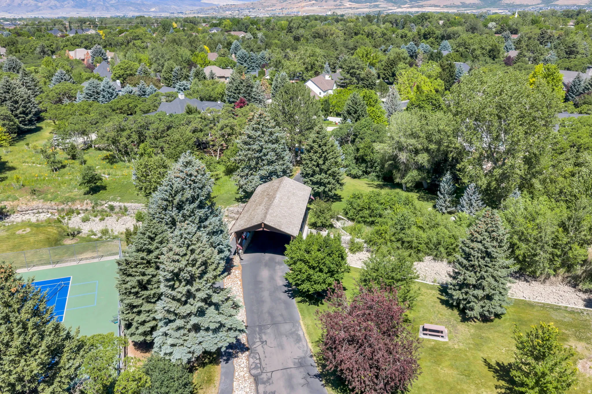 Expansive property featuring a private tennis court, covered parking structure, paved driveway, mature trees, and a mountain backdrop