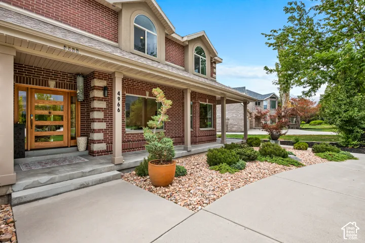 Brick facade with stone accents, covered entry, wood-finish entry door with clear glass panels, arched dormer window, and mature landscaping