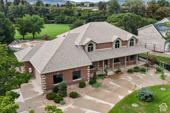 Brick exterior with multiple dormer windows, covered front porch, concrete driveway, attached garage, and an in-ground pool