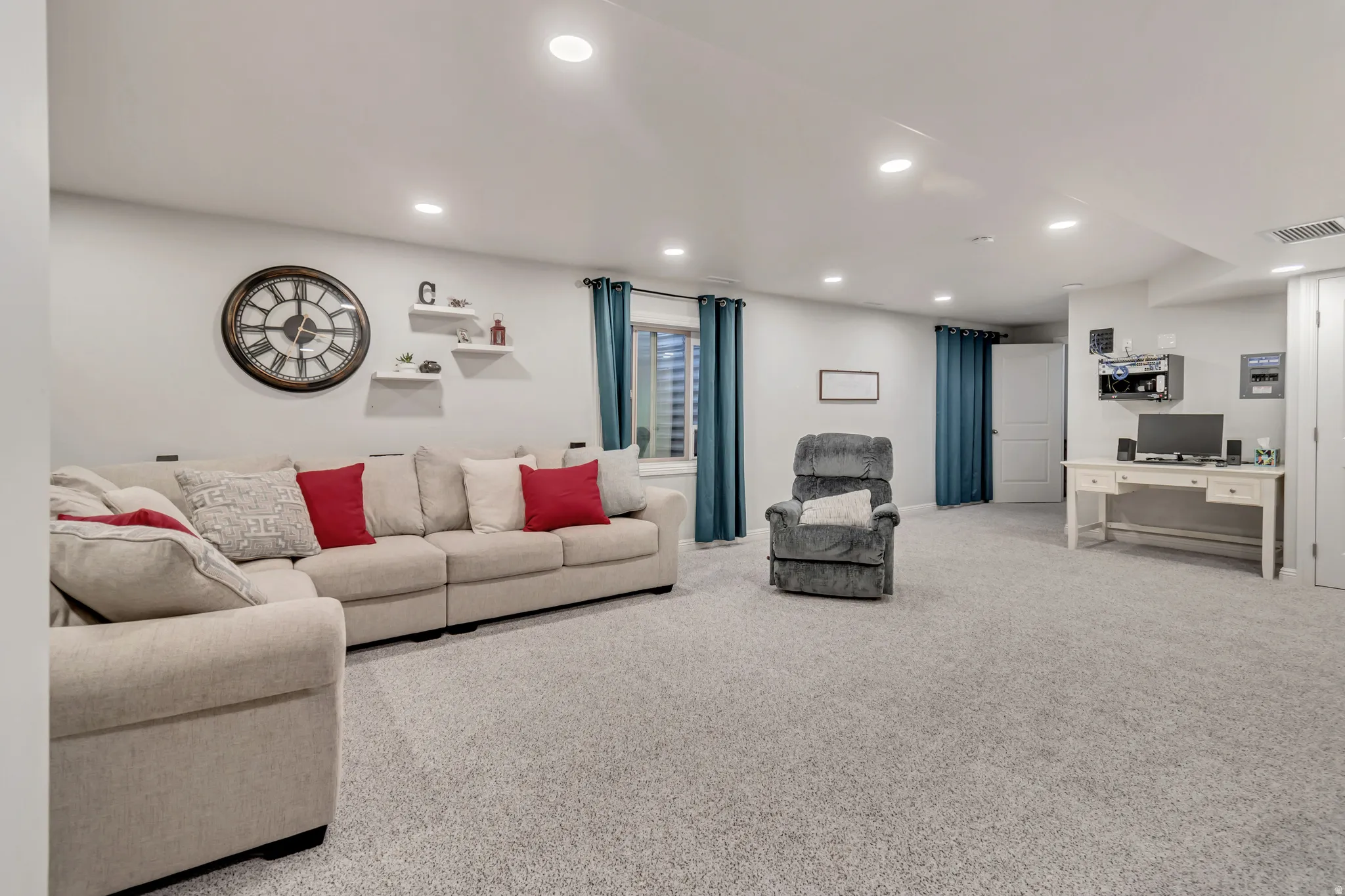 Spacious carpeted room featuring recessed lighting, multiple windows with teal curtains, built-in floating shelves, and a white paneled door